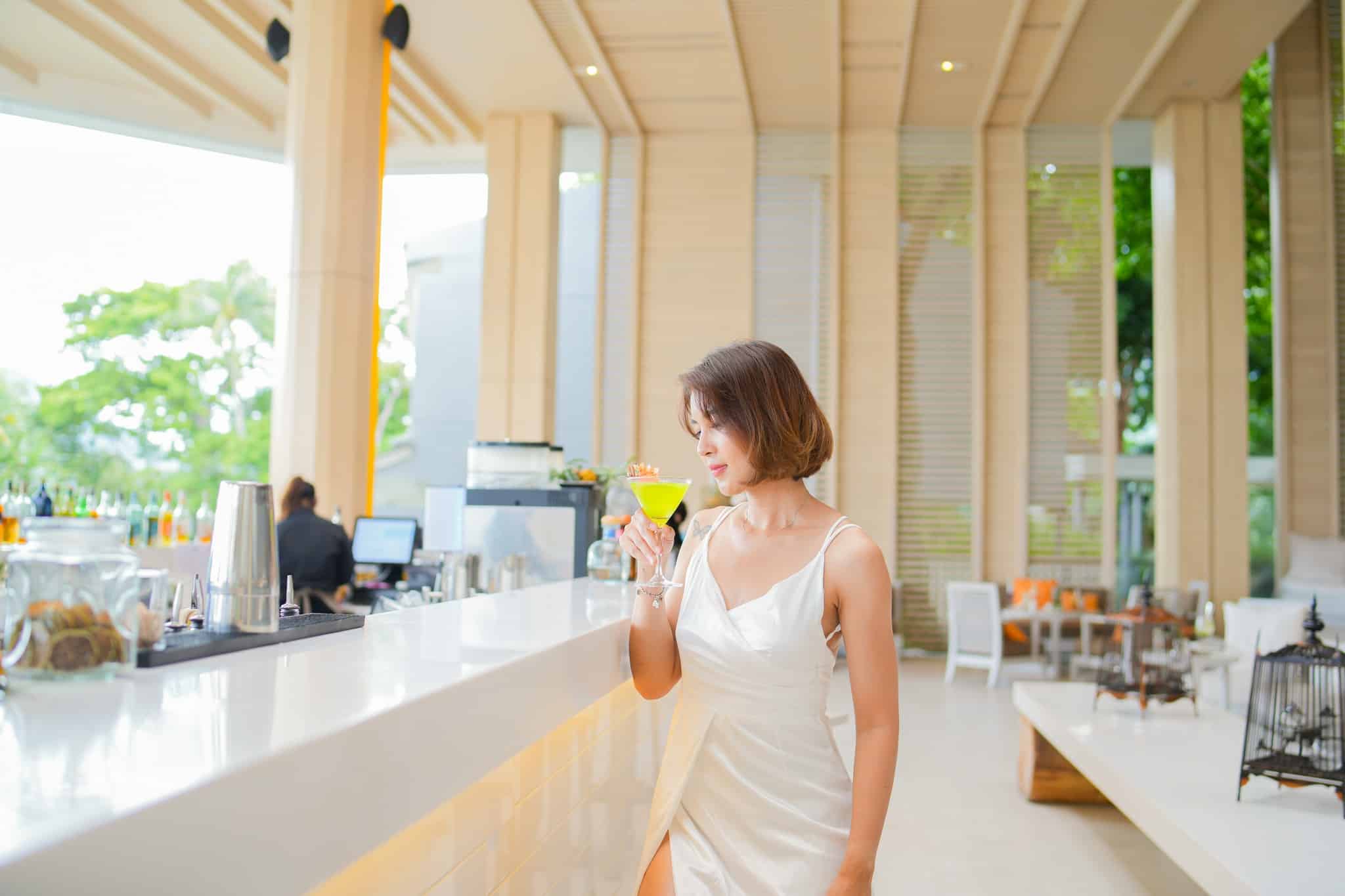 ผู้หญิงในชุดเดรสสีขาว ยืนอยู่ที่บาร์สมัยใหม่ พร้อมถือค็อกเทลสีเหลืองไว้ในมือ โดยมีแสงสว่างจากธรรมชาติที่สดใสเติมเต็มบรรยากาศ—เหมาะสำหรับการบันทึกช่วงเวลาที่เก๋ไก๋ที่ ที่เที่ยวป่าตอง.