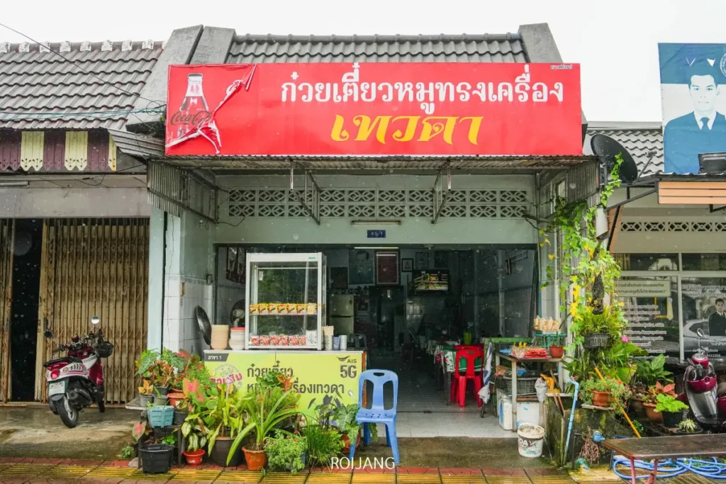 ภาพถนนหน้าร้าน ขนมจีนป้าม่อม ร้านก๋วยเตี๋ยวไทยขนาดเล็ก มีป้ายสีแดง ต้นไม้กระถางวางอยู่หน้าร้าน และมองเห็นเก้าอี้และโต๊ะพลาสติกด้านใน.