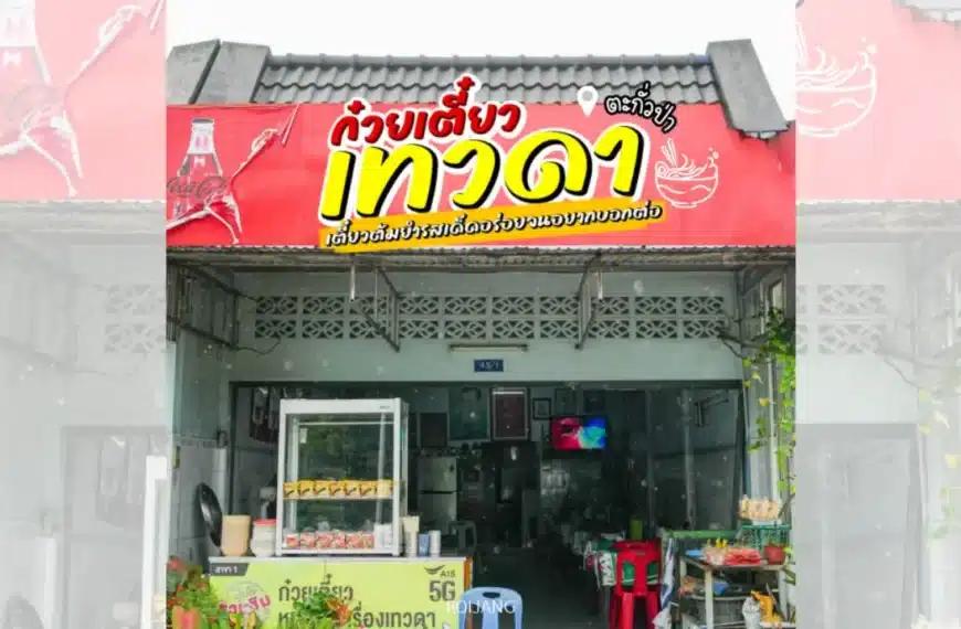 ร้านก๋วยเตี๋ยวไทยขนาดเล็ก มีป้ายสีแดงพร้อมตัวอักษรไทยสีขาวและเหลือง ชื่อร้านว่า ก๋วยเตี๋ยวเทวดา มีภาพประกอบไก่ตัวผู้ และโลโก้ Coca-Cola อยู่เหนือทางเข้าที่เปิดอยู่.
