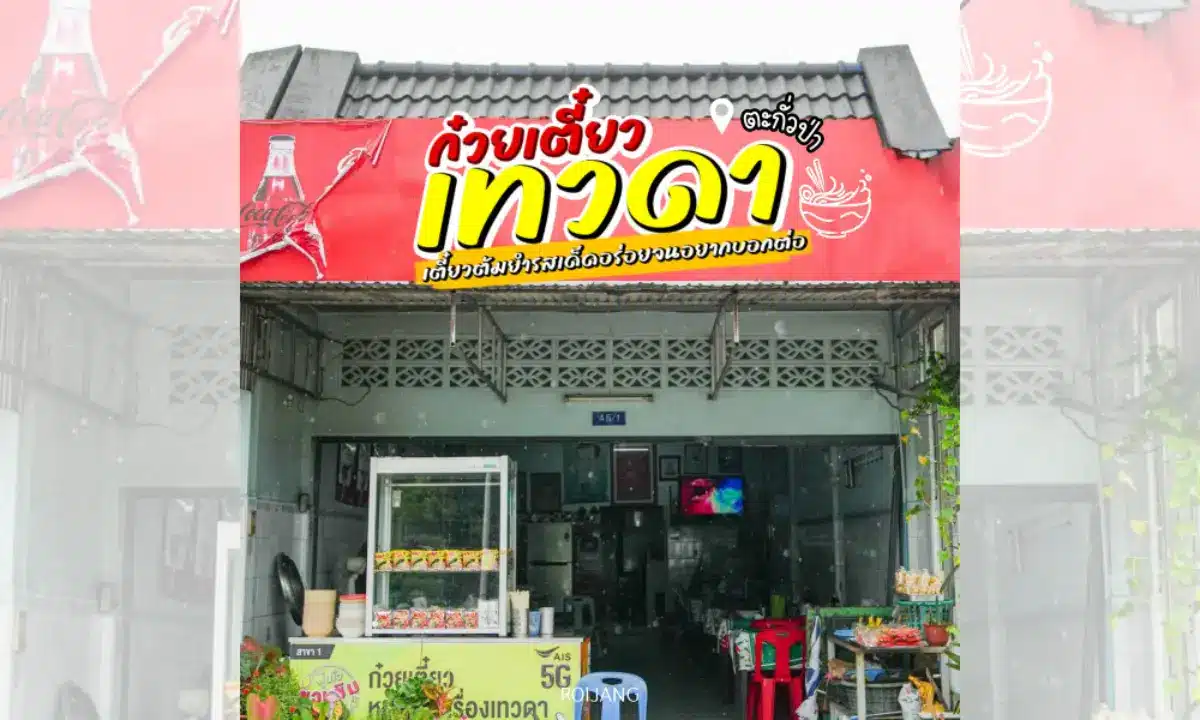 ร้านก๋วยเตี๋ยวไทยขนาดเล็ก มีป้ายสีแดงพร้อมตัวอักษรไทยสีขาวและเหลือง ชื่อร้านว่า ก๋วยเตี๋ยวเทวดา มีภาพประกอบไก่ตัวผู้ และโลโก้ Coca-Cola อยู่เหนือทางเข้าที่เปิดอยู่.