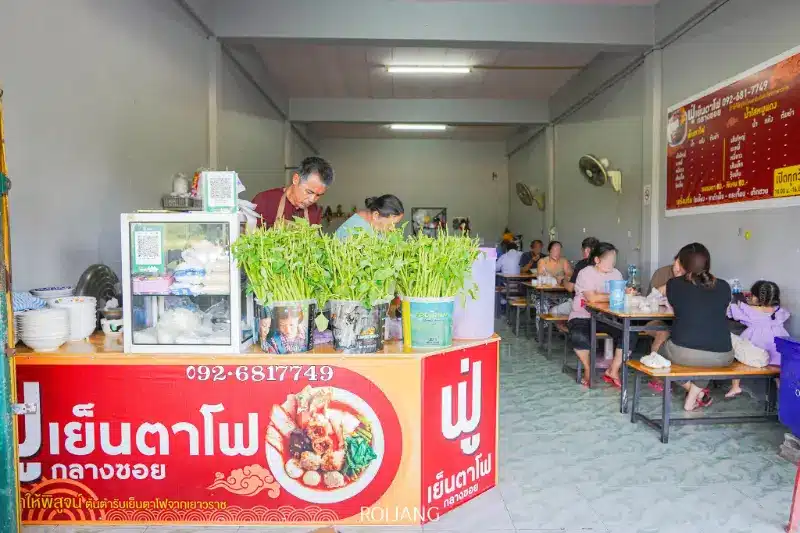 ภายในร้านอาหารขนาดเล็กที่มีผู้คนนั่งรับประทานอาหารที่โต๊ะไม้; ผู้ขายเตรียมฟูเย็นตาโฟอยู่หลังเคาน์เตอร์ที่มีป้ายสีแดงและสมุนไพรสดวางอยู่ด้านหน้า.