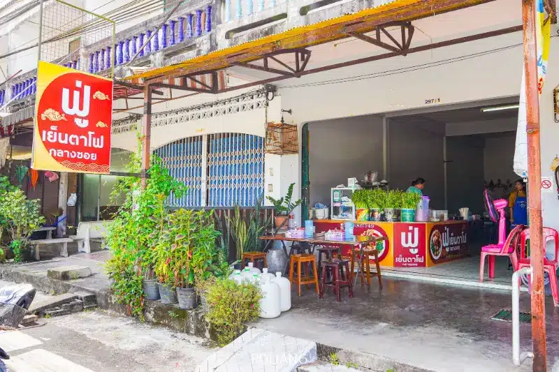 ร้านอาหารไทยเปิดโล่งด้านหน้า มีป้ายสีแดง ต้นไม้กระถางด้านนอก เก้าอี้และโต๊ะพลาสติก พร้อมเคาน์เตอร์ที่ให้บริการฟูเย็นตาโฟและอาหารและเครื่องดื่มภายในร้าน.