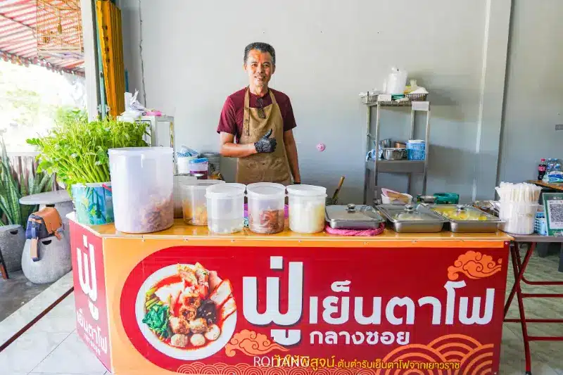 ชายคนหนึ่งสวมผ้ากันเปื้อนและถุงมือ ยืนอยู่หลังเคาน์เตอร์ร้านขายอาหาร มีภาชนะบรรจุส่วนผสมต่าง ๆ พร้อมเสิร์ฟเมนูบะหมี่ไทย เช่น ฟู่เย็นตาโฟ เคาน์เตอร์มีป้ายสีแดงพร้อมข้อความภาษาไทยและภาพอาหารที่ชวนให้ลิ้มลอง.