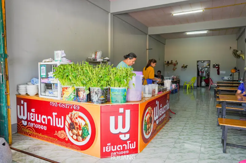 ร้านอาหารไทยขนาดเล็กที่มีเคาน์เตอร์สีแดง, ต้นไม้กระถาง, จานซ้อนกัน, และผู้คนกำลังเตรียมฟูเย็นตาโฟ; มีโต๊ะและเก้าอี้ไม้ในห้องอาหาร.