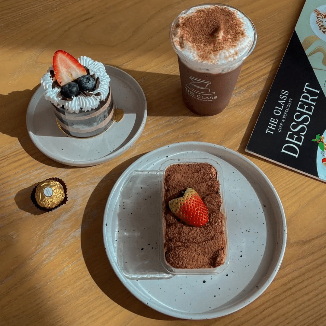 โต๊ะที่มีเค้กช็อกโกแลตชั้น, ทีรามิสุที่โรยด้วยสตรอเบอร์รี่, เครื่องดื่ม ที่เที่ยวชุมพร ช็อกโกแลตฟองนุ่ม, เฟอเรโร่ โรเช่, และเมนูของหวาน—เหมาะสำหรับการผ่อนคลายหลังจากสำรวจ ชุมพรที่เที่ยว.