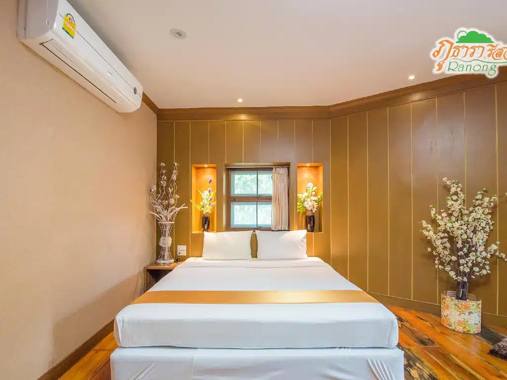 เตียงที่จัดไว้อย่างเรียบร้อยด้วยผ้าปูที่นอนสีขาวและทองตั้งอยู่ตรงกลางห้องที่ผนังเป็นไม้ที่ โรงแรมระนอง ขนาบข้างด้วยแจกันประดับและหน้าต่างบานเล็ก เครื่องปรับอากาศติดตั้งอยู่บนผนัง.