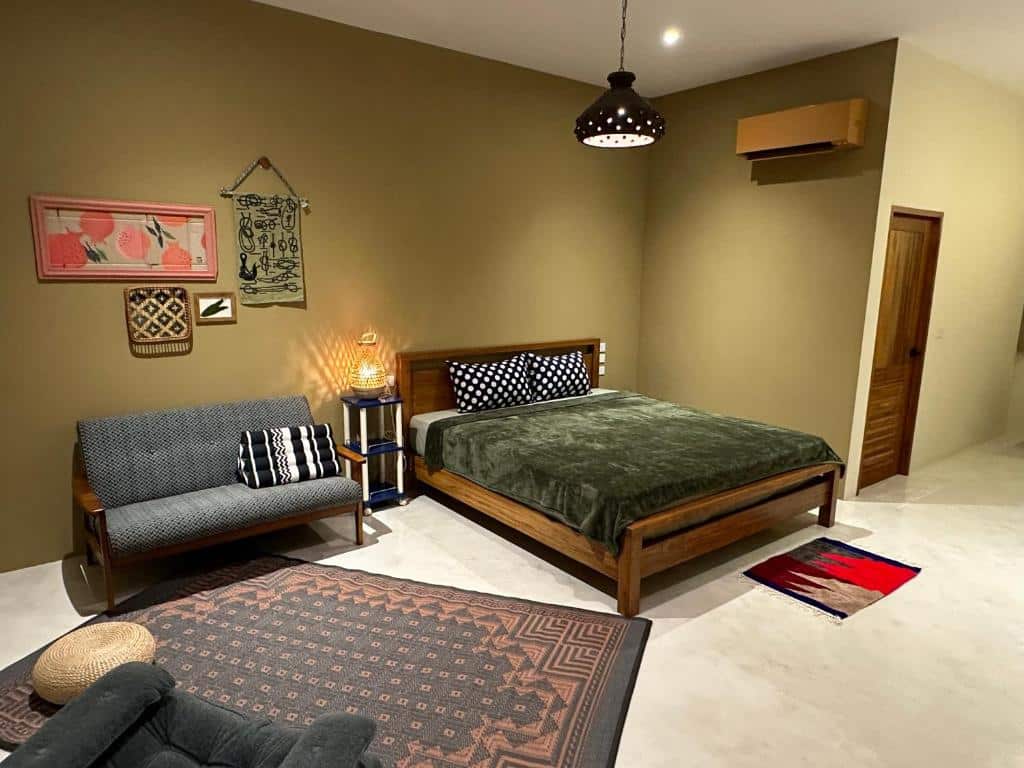 ห้องนอนที่อบอุ่นที่ ที่พักติดทะเลปราณบุรี ประกอบด้วยเตียงไม้พร้อมผ้าห่มสีเขียว, โซฟาขนาดเล็ก, ภาพศิลปะบนผนัง, พรมลาย, โคมไฟ, และประตูไม้ปิดบนผนังสีเบจและพื้นสีขาว.