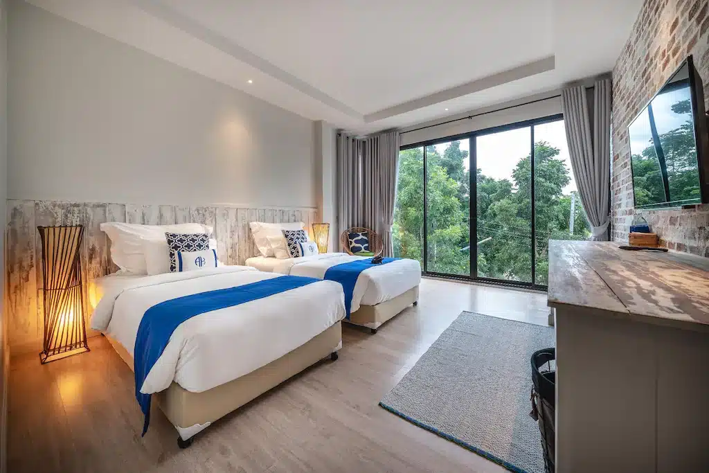 ห้องพักโรงแรมสมัยใหม่พร้อมเตียงเดี่ยวสองเตียง ตกแต่งด้วยสีน้ำเงิน หน้าต่างบานใหญ่ที่มองเห็นวิวสีเขียว พื้นไม้ และการตกแต่งร่วมสมัย—เหมาะสำหรับผู้ที่มองหาที่พักติดทะเลในปราณบุรีราคาไม่แพงสำหรับการพักผ่อนครั้งต่อไปของคุณ.
