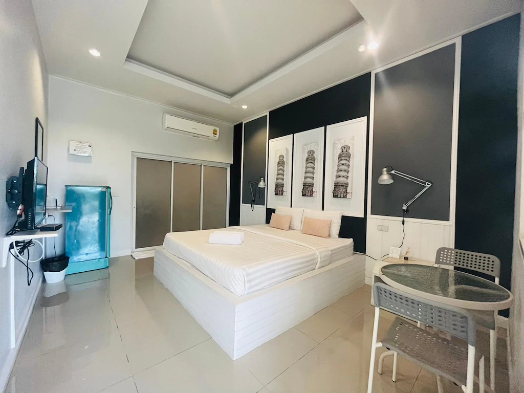 ห้องนอนสไตล์โมเดิร์นมินิมอลที่ ที่พักปราณบุรีติดทะเลเป็นหลัง ประกอบด้วยเตียงคู่, งานศิลปะบนผนัง, โต๊ะรับประทานอาหารสำหรับสองคน, ตู้เย็นสีฟ้า, โทรทัศน์, เครื่องปรับอากาศ, การตกแต่งสีขาวดำ, และพื้นกระเบื้อง—เหมาะสำหรับการพักผ่อนริมทะเลอย่างมีสไตล์ของคุณ.