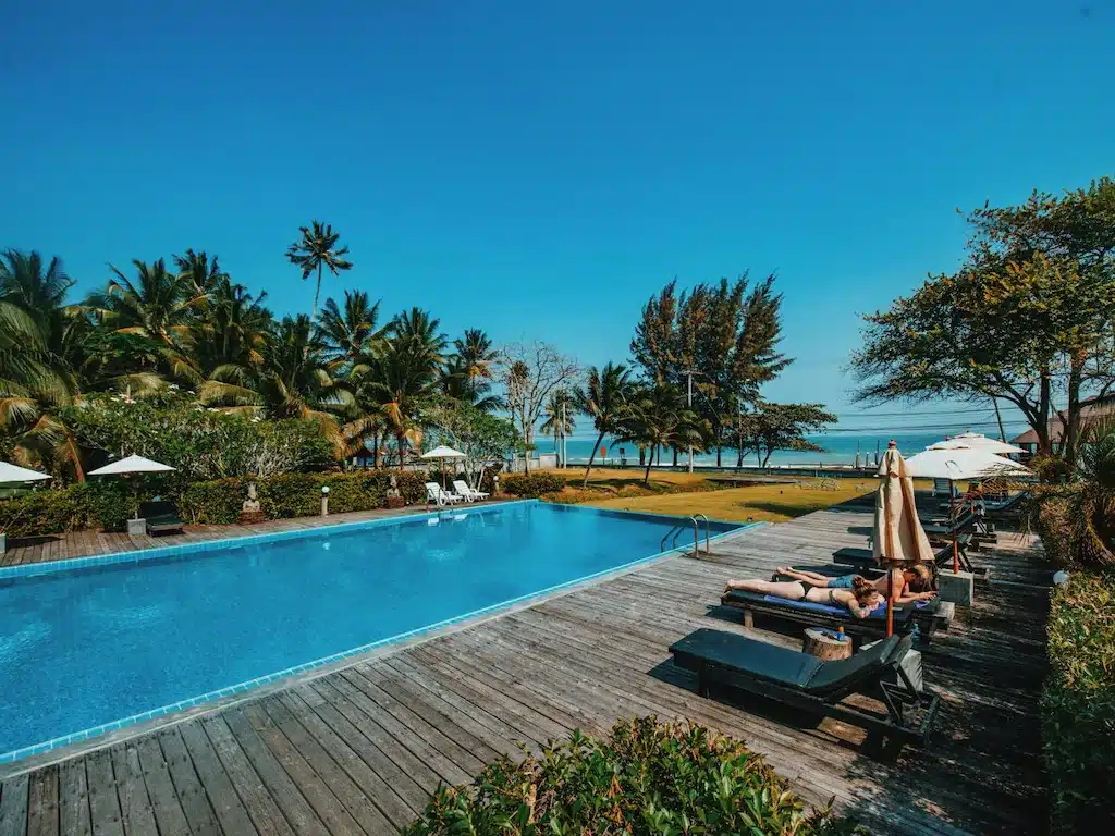 สระว่ายน้ำกลางแจ้งพร้อมเก้าอี้อาบแดดบนดาดฟ้าไม้ ล้อมรอบด้วยต้นปาล์ม ให้ทัศนียภาพที่สวยงาม ที่เที่ยวชุมพร ของชายหาดและทะเลภายใต้ท้องฟ้าสีครามใส—เหมาะสำหรับการผ่อนคลายขณะสำรวจสถานที่ท่องเที่ยวในชุมพร.