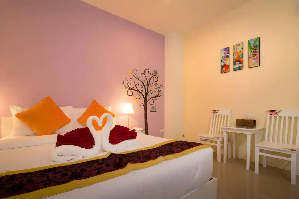  ที่พักระนอง เตียงที่จัดไว้อย่างเรียบร้อยพร้อมผ้าเช็ดตัวรูปหงส์และตกแต่งด้วยสีแดง เก้าอี้สีขาวสองตัวกับโต๊ะเล็ก ๆ ผนังสีม่วงพาสเทล และงานศิลปะบนผนังสีสันสดใสในโรงแรมระนองที่สว่างไสวเพื่อการพักผ่อนในเมืองที่แสนอบอุ่น.