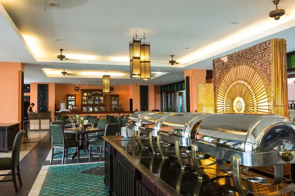 พื้นที่บุฟเฟ่ต์ของร้านอาหารที่มีจานเสิร์ฟแบบปิดบนเคาน์เตอร์ โต๊ะและเก้าอี้สำหรับรับประทานอาหาร แสงไฟอบอุ่น และฉากตกแต่งในพื้นหลัง—เหมาะอย่างยิ่งสำหรับแขกที่ต้องการเพลิดเพลินกับประสบการณ์การพักผ่อนที่ที่พักติดทะเลในปราณบุรี.
