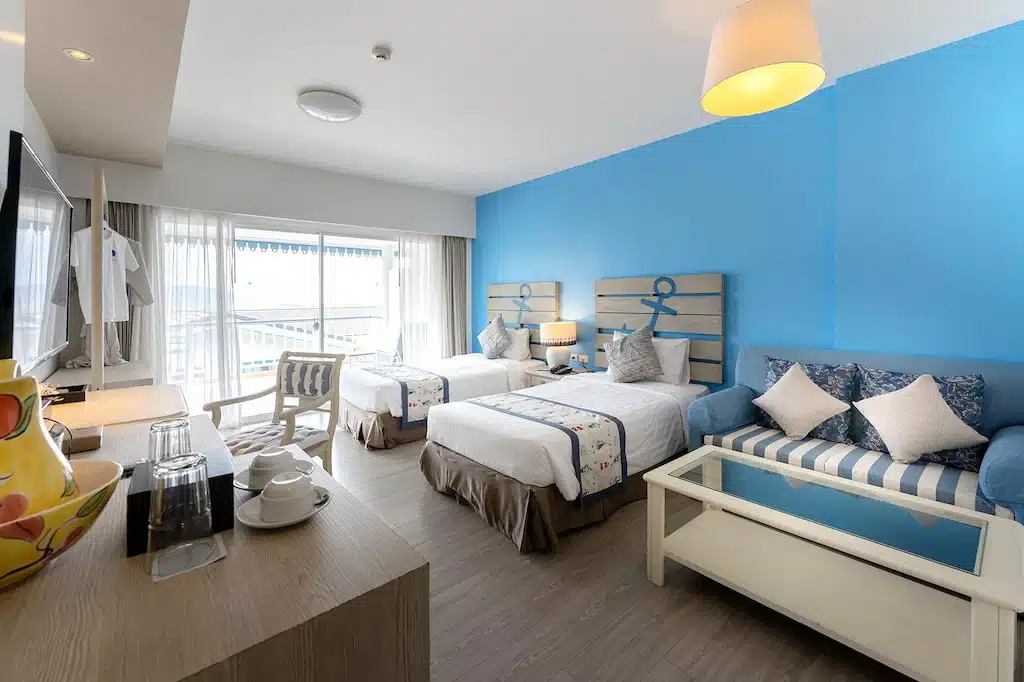 ห้องพักโรงแรมพร้อมเตียงเดี่ยวสองเตียง ผนังสีน้ำเงินเป็นจุดเด่น การตกแต่งสไตล์ทะเล โซฟาลายทาง รวม ที่เที่ยวเกาะสีชัง เกาะสวยๆ ในที่เที่ยวชลบุรี ไป เที่ยวเกาะสีชัง เดินทางง่ายๆ ใกล้กรุงเทพแบบไปเช้าเย็นกลับได้ หลีกหนีความวุ่นวาย พร้อมอวดสถานที่สโลว์ไลฟ์
และโต๊ะกาแฟกระจก—เหมาะสำหรับการพักผ่อนหลังจากเที่ยวเกาะสีชังมาทั้งวัน หน้าต่างบานใหญ่พร้อมผ้าม่านบางช่วยให้แสงธรรมชาติส่องเข้ามาได้อย่างเต็มที่.