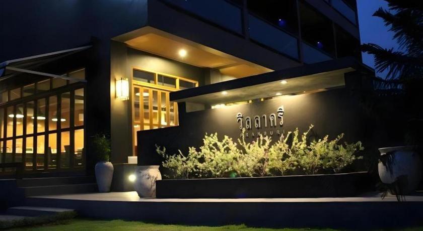 อาคารภายนอกสมัยใหม่ในยามค่ำคืนพร้อมป้ายไฟที่เขียนด้วยอักษรไทย หน้าต่างบานใหญ่ พืชภายนอกอาคาร และแจกันประดับตกแต่ง—เหมาะสำหรับผู้ที่มองหา ที่พักติดทะเลปราณบุรี ในบรรยากาศที่มีสไตล์.