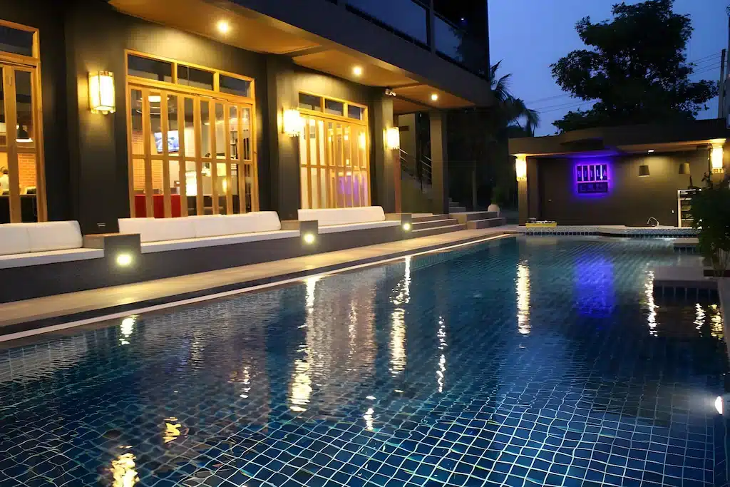 สระว่ายน้ำกลางแจ้งสมัยใหม่ในยามค่ำคืน ล้อมรอบด้วยที่นั่งสีขาวและไฟผนังอ่อนโยน สะท้อนให้เห็นถึงภายนอกของอาคารและต้นไม้ใกล้เคียง—เหมาะอย่างยิ่งสำหรับการพักผ่อนที่ ที่พักปราณบุรีติดทะเลเป็นหลัง.