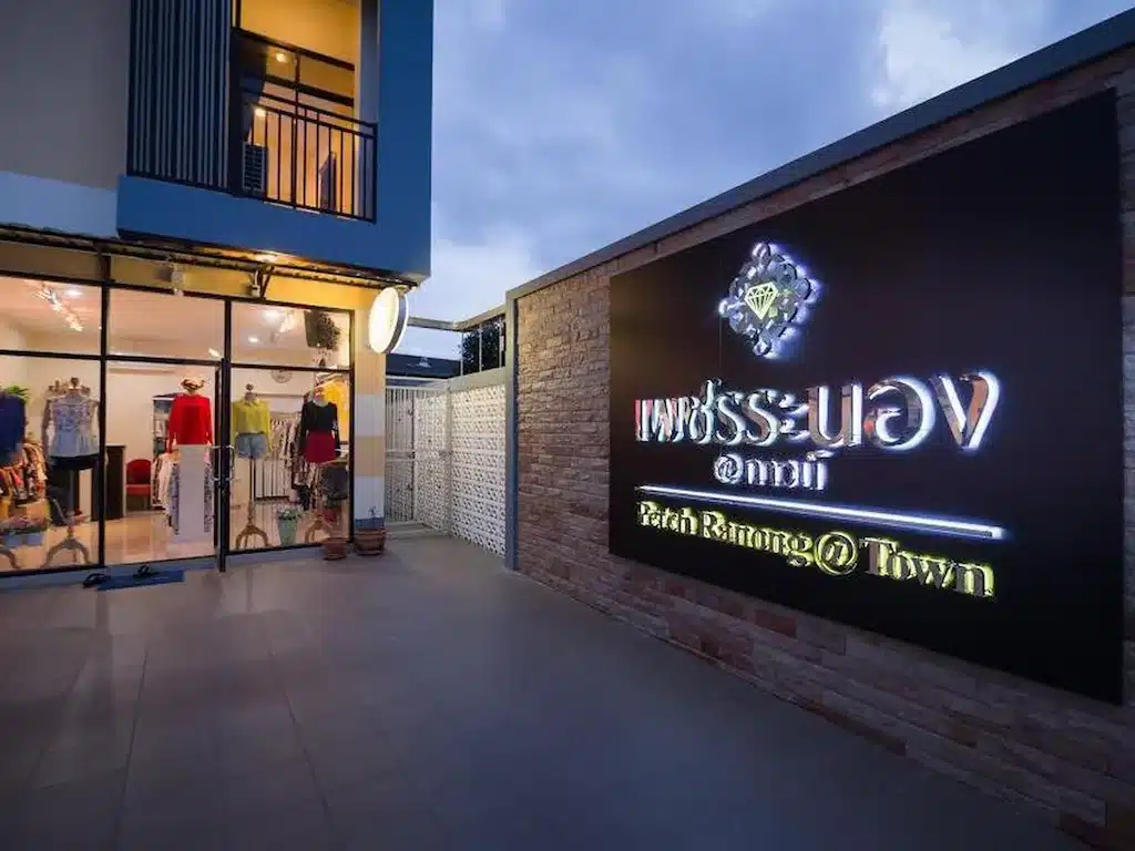 ทางเข้าของร้านเสื้อผ้าที่มีหน้าต่างกระจกโชว์หุ่นโชว์ตัว และป้ายไฟ ที่พักระนอง ขนาดใหญ่ที่เขียนว่า "Patch Ranong @ Town" ทั้งภาษาอังกฤษและภาษาไทย ตั้งอยู่ใกล้ที่พักระนองในเมือง สะดวกสบายสำหรับการช้อปปิ้งหลังจากที่คุณเข้าพัก.