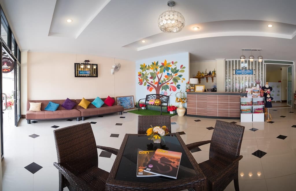 ล็อบบี้โรงแรมสมัยใหม่ที่ โรงแรมระนอง ประกอบด้วยเคาน์เตอร์ต้อนรับ, ภาพจิตรกรรมฝาผนังต้นไม้, พื้นที่นั่งเล่นพร้อมหมอนสีสันสดใส และโต๊ะพร้อมนิตยสารและดอกไม้จัดเล็ก ๆ—เหมาะสำหรับแขกที่กำลังมองหาที่พักในเมืองระนองที่มีสไตล์.