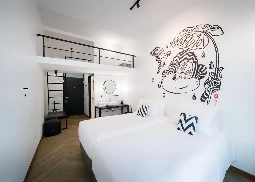 ห้องพัก โรงแรมระนอง สมัยใหม่พร้อมเตียงคู่สองเตียง ชุดเครื่องนอนสีขาว ศิลปะติดผนังขาวดำ พื้นที่ล้างหน้าขนาดเล็ก และพื้นที่ลอฟท์เหนือทางเข้า—เหมาะสำหรับผู้ที่มองหา โรงแรมระนอง ใจกลางเมือง.