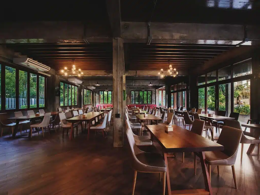 ร้านอาหารสมัยใหม่ ว่างเปล่า พร้อมพื้นไม้ หน้าต่างขนาดใหญ่ โต๊ะไม้หลายตัว และเก้าอี้สีเบจ ส่องสว่าง ที่เที่ยวชุมพร ด้วยแสงธรรมชาติและโคมไฟเพดาน—สถานที่เชิญชวนให้ผ่อนคลายขณะสำรวจสถานที่ท่องเที่ยวในชุมพร.