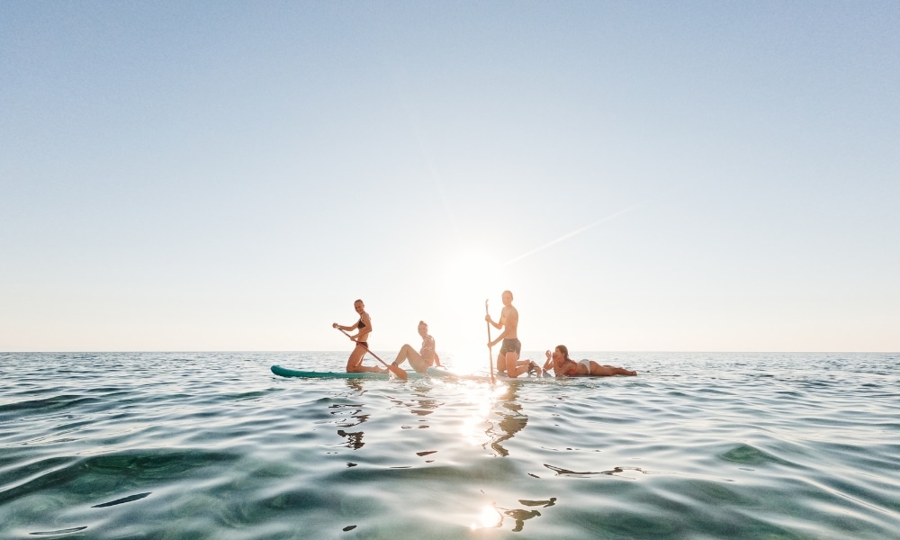 ห้าคนกำลังเล่นแพดเดิลบอร์ดบนผืนน้ำที่สงบในระหว่างวันที่แดดจ้า ดื่มด่ำกับทัศนียภาพอันน่าทึ่งที่หนึ่งในสถานที่ท่องเที่ยวชุมพรยอดนิยม โดยมีดวงอาทิตย์กำลังตกต่ำลงที่ ที่เที่ยวชุมพร ขอบฟ้าและท้องฟ้าที่แจ่มใสอยู่เบื้องบน.