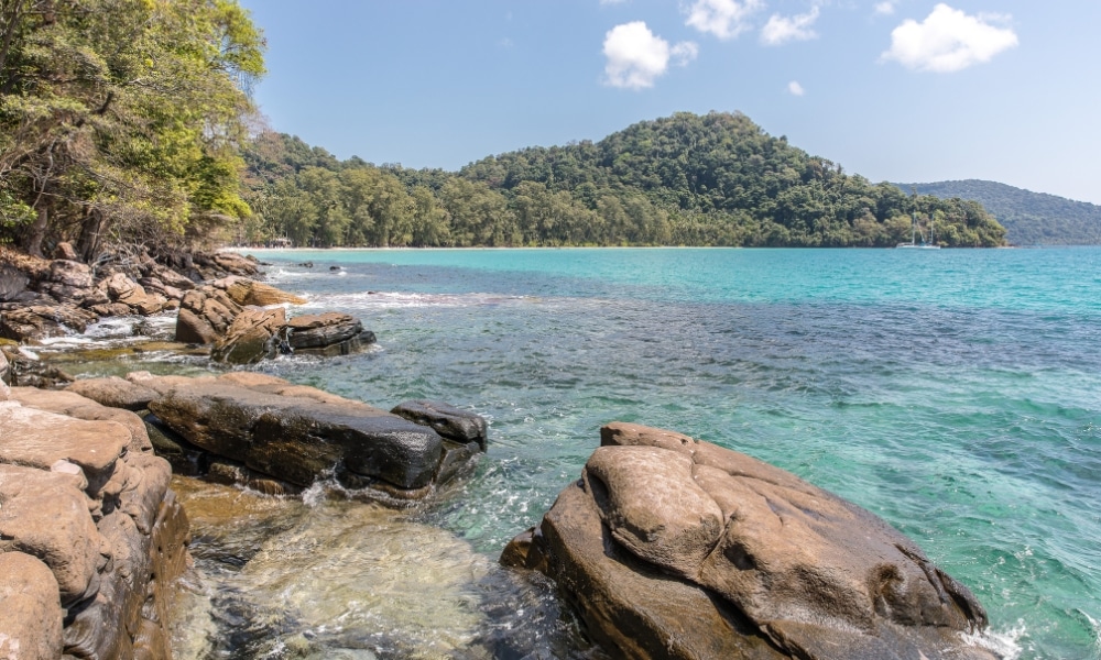 ชายฝั่งหินที่มีน้ำทะเลใสสีเทอร์ควอยซ์และภูเขาที่ปกคลุมไปด้วยป่าไม้ในฉากหลังภายใต้ท้องฟ้าสีครามรวม ที่เที่ยวเกาะสีชัง เกาะสวยๆ ในที่เที่ยวชลบุรี ไป เที่ยวเกาะสีชัง เดินทางง่ายๆ ใกล้กรุงเทพแบบไปเช้าเย็นกลับได้ หลีกหนีความวุ่นวาย พร้อมอวดสถานที่สโลว์ไลฟ์
ที่มีเมฆลอยอยู่เป็นระยะ—เหมาะสำหรับผู้ที่ต้องการ เที่ยวเกาะสีชัง และค้นพบความงามตามธรรมชาติของที่นี่.