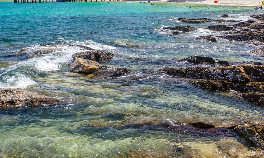 น้ำทะเลสีฟ้าอมเขียวใสไหลผ่านชายฝั่งที่เต็มไปด้วยโขดหินพร้อมคลื่นเล็ก ๆ และฟองคลื่น; หาดทรายรวม ที่เที่ยวเกาะสีชัง เกาะสวยๆ ในที่เที่ยวชลบุรี ไป เที่ยวเกาะสีชัง เดินทางง่ายๆ ใกล้กรุงเทพแบบไปเช้าเย็นกลับได้ หลีกหนีความวุ่นวาย พร้อมอวดสถานที่สโลว์ไลฟ์
และท่าเรือสามารถมองเห็นได้ในพื้นหลัง—เหมาะสำหรับผู้ที่ต้องการไปเที่ยวเกาะสีชังและสำรวจความสวยงามของทิวทัศน์.