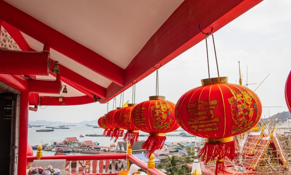 โคมไฟจีนสีแดงแขวนอยู่จากหลังคาพร้อมการตกแต่งสีทอง มองเห็นท่าเรือที่มีเรือและอาคารในระยะไกล—รวม ที่เที่ยวเกาะสีชัง เกาะสวยๆ ในที่เที่ยวชลบุรี ไป เที่ยวเกาะสีชัง เดินทางง่ายๆ ใกล้กรุงเทพแบบไปเช้าเย็นกลับได้ หลีกหนีความวุ่นวาย พร้อมอวดสถานที่สโลว์ไลฟ์
ถ่ายทอดเสน่ห์อันเป็นเอกลักษณ์ของ ที่เที่ยวเกาะสีชัง.