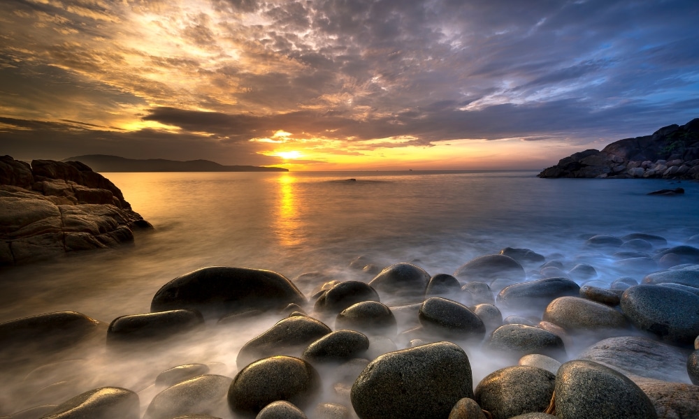 ชายฝั่งหินในยามพระอาทิตย์ตกดิน ณ ที่เที่ยวเกาะสีชัง, พร้อมคลื่นน้ำรวม ที่เที่ยวเกาะสีชัง เกาะสวยๆ ในที่เที่ยวชลบุรี ไป เที่ยวเกาะสีชัง เดินทางง่ายๆ ใกล้กรุงเทพแบบไปเช้าเย็นกลับได้ หลีกหนีความวุ่นวาย พร้อมอวดสถานที่สโลว์ไลฟ์
ทะเลที่นุ่มนวลและหมอกบางในเบื้องหน้า และท้องฟ้าที่มีเมฆปกคลุมซึ่งถูกส่องสว่างโดยพระอาทิตย์ที่กำลังลับขอบฟ้าในเบื้องหลัง.