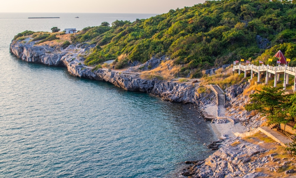 ชายฝั่งที่เต็มไปด้วยโขดหินและพืชพรรณสีเขียว มีทางเดินและศาลาพร้อมธงปลิวไสวเหนือผืนน้ำสีฟ้าสงบภายใต้ท้องฟ้าแจ่มใส—ภาพที่ชวนให้ใครก็ตามที่ต้องการ เที่ยวเกาะสีชัง และเพลิดเพลินกับความงามของธรรมชาติ.