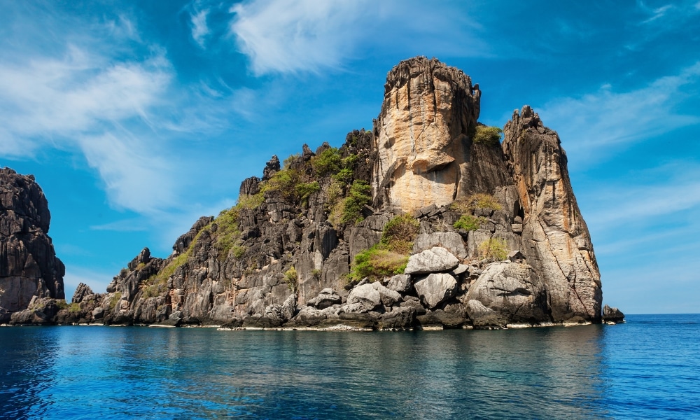 เกาะหินปูนสีเทาสูงตระหง่านขึ้นมาจากน้ำทะเลใสสีฟ้าภายใต้ท้องฟ้าที่มีเมฆบางส่วนปกคลุม มีพืชพรรณที่กระจัดกระจายอยู่บนหน้าผาที่ขรุขระ—เป็นอัญมณีที่ซ่อนอยู่ท่ามกลางสถานที่ท่องเที่ยวในชุมพรสำหรับผู้ที่ต้องการสำรวจความงามตามธรรมชาติของ สถานที่ท่องเที่ยวในชุมพร.