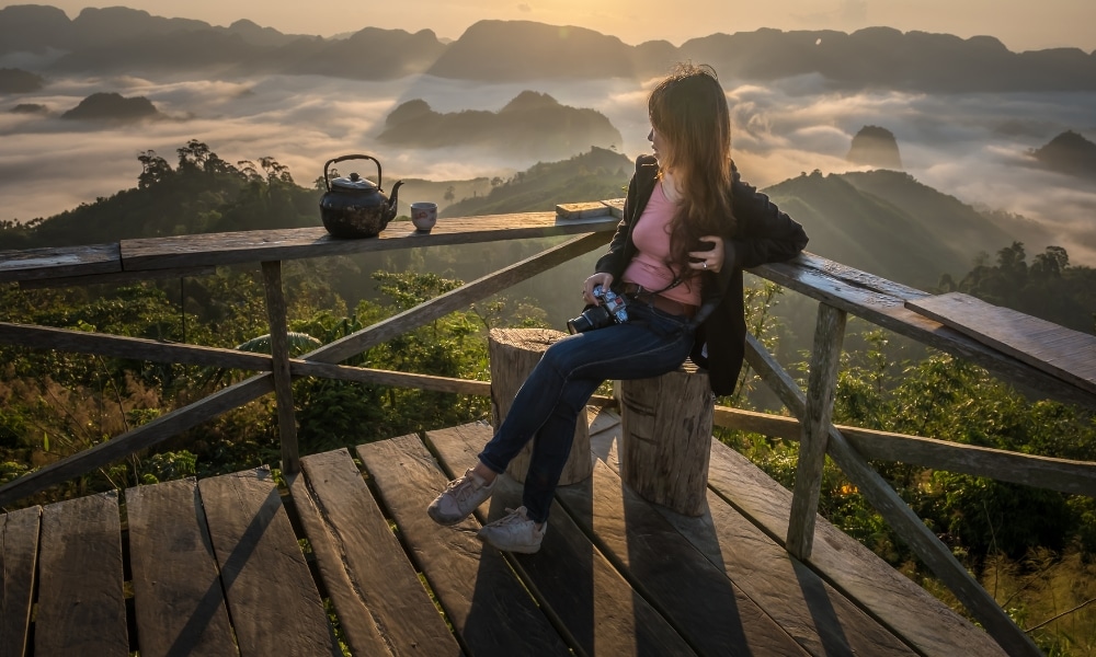 ผู้หญิงคนหนึ่งนั่งอยู่บนดาดฟ้าไม้ที่ ที่เที่ยวชุมพร มองเห็นภูเขาหมอกยามพระอาทิตย์ขึ้น ถือกล้องถ่ายรูปไว้ มีกาน้ำชาและถ้วยอยู่ใกล้ๆ กำลังเพลิดเพลินกับความงดงามอันเงียบสงบของ ชุมพรที่เที่ยว.
