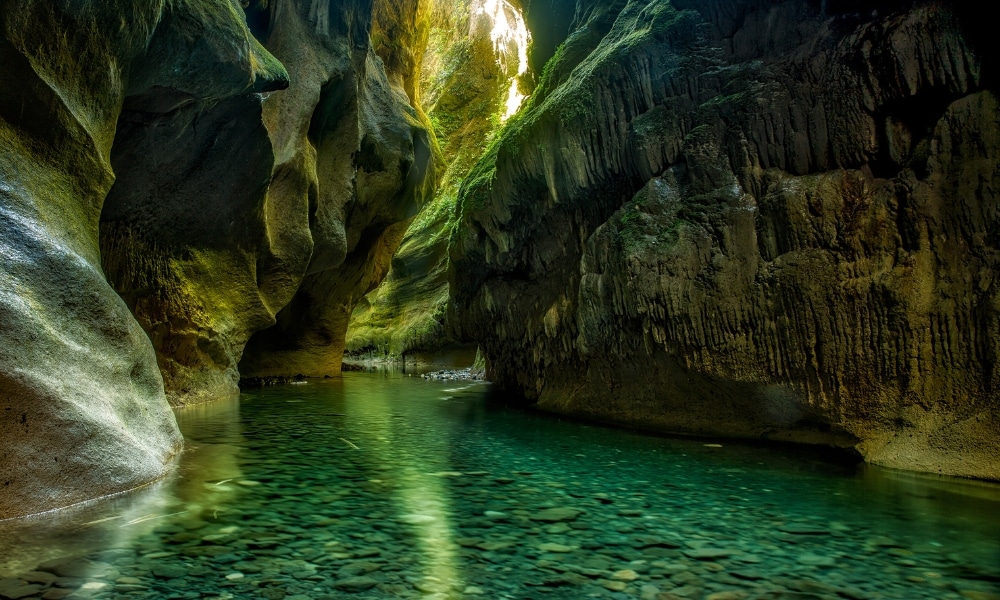 ลำธารใสไหลผ่านหุบเขาแคบและ ที่เที่ยวชุมพร ขรุขระที่มีผนังสูงชันปกคลุมด้วยมอสและแสงแดดที่ส่องลงมา—ภาพที่งดงามราวกับสวรรค์ในสถานที่ท่องเที่ยวที่ซ่อนอยู่ในชุมพรสำหรับผู้รักธรรมชาติ.