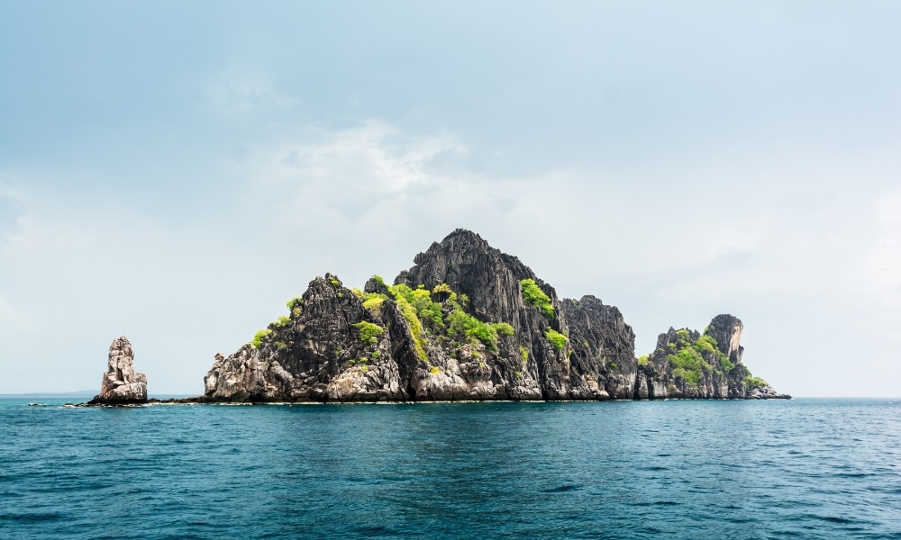 เกาะหินที่มีพืชสีเขียวขึ้นอยู่อย่าง ที่เที่ยวชุมพร เบาบางโผล่ขึ้นมาจากมหาสมุทรสีครามภายใต้ท้องฟ้าที่มีเมฆปกคลุม—ภาพอันเป็นเอกลักษณ์สำหรับผู้ที่มองหาสถานที่ท่องเที่ยวในชุมพรตามแนวชายฝั่งที่สวยงามตระการตา.