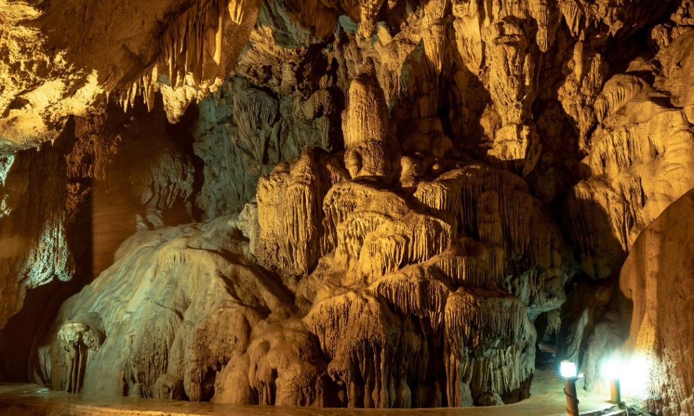 บุคคลหนึ่งสวมไฟฉายคาดศีรษะยืนอยู่ภายในถ้ำขนาดใหญ่ใน ชุมพรที่เที่ยว ล้อมรอบด้วยหินงอกหินย้อยที่ส่องแสงระยิบระยับและหินงอกที่สวยงามตระการตา.