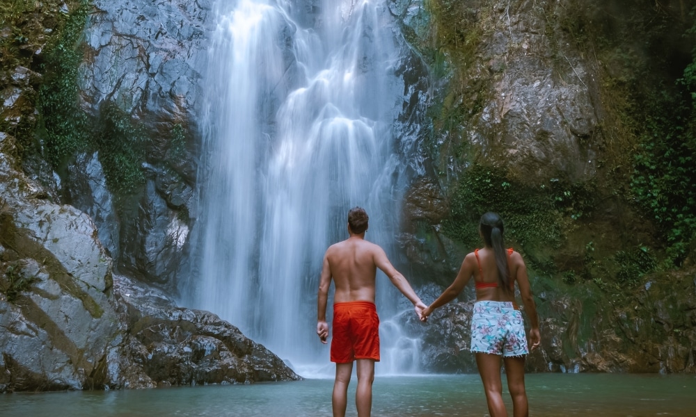 ชายและหญิงจับมือกันยืนอยู่หน้า น้ำตกขนาดใหญ่ ในบรรยากาศที่เต็มไปด้วย ที่เที่ยวชุมพร หินและพืชพรรณเขียวชอุ่ม ค้นพบหนึ่งในสถานที่ท่องเที่ยวที่สวยงามของชุมพร.