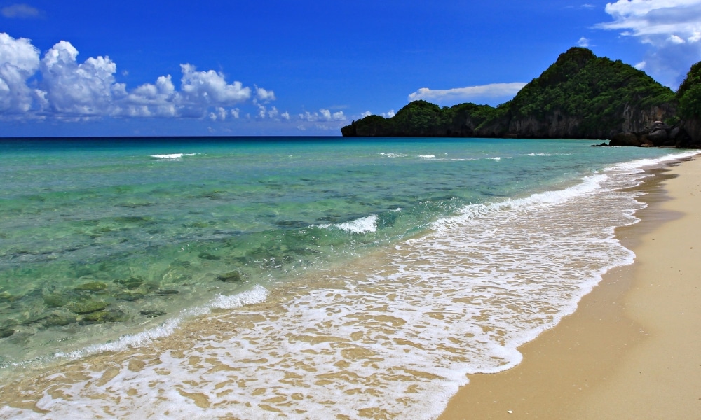 คลื่นสีฟ้าใสกระทบ เที่ยวชุมพร ชายหาดทรายพร้อมฉากหลังเป็นเนินเขาสีเขียวและท้องฟ้าสีครามที่มีเมฆลอยเป็นจุด ๆ—เหมาะสำหรับผู้ที่กำลังมองหาสถานที่ท่องเที่ยวในชุมพร.