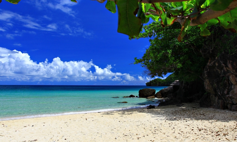 ชายหาดทรายขาวสะอาด น้ำทะเลใสสีเทอร์ควอยซ์ กระจายไปด้วยก้อนหิน ต้นไม้เขียวชอุ่ม และท้องฟ้าสีฟ้า ที่เที่ยวชุมพร สดใสพร้อมเมฆขาว—หนึ่งในสถานที่ท่องเที่ยวที่สวยที่สุดของชุมพรสำหรับผู้รักธรรมชาติ.