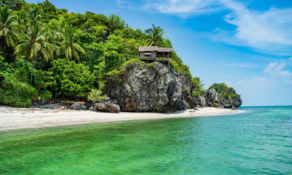 กระท่อมไม้หลังหนึ่งตั้งอยู่บนหน้าผาหิน ล้อมรอบด้วยต้นไม้เขตร้อนหนาแน่น มองเห็นชายหาดทรายและ เที่ยวชุมพร ทะเลสีเขียวอมฟ้าใสภายใต้ท้องฟ้าที่มีเมฆบางส่วน—เป็นภาพที่งดงามราวกับฝันในสถานที่ท่องเที่ยวที่ดีที่สุดในชุมพร.