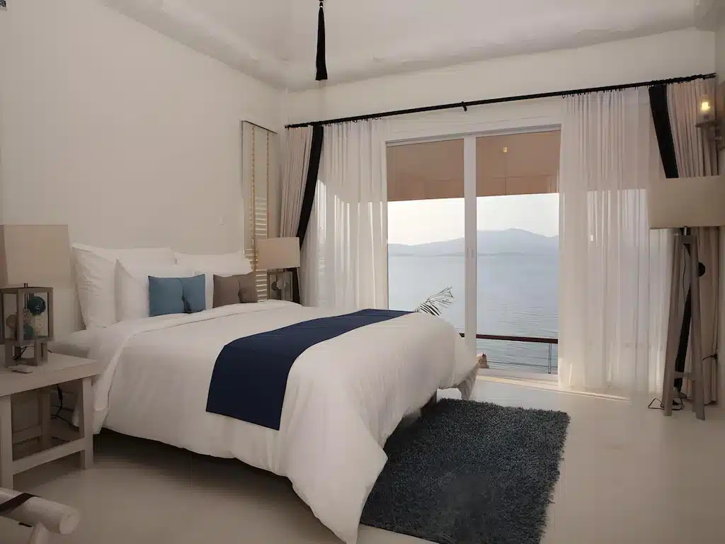 ห้องนอนสไตล์โมเดิร์นพร้อมเตียงขนาดใหญ่ ตกแต่งด้วยสีน้ำเงิน พรมสีเทา และหน้าต่างบานใหญ่จากพื้นจรดเพดานที่มองเห็นทะเลและภูเขา—เหมาะสำหรับผู้ที่ต้องการที่พักในเมืองระนองพร้อมวิวที่สวยงาม ที่พักระนอง ตระการตา.