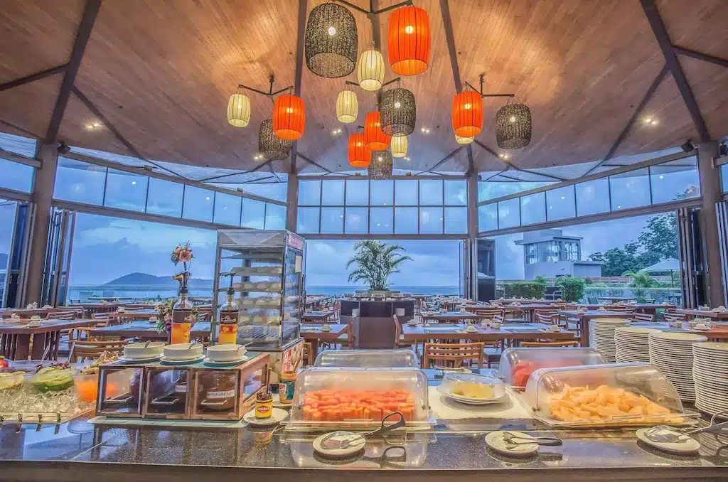 ร้านอาหารกว้างขวางพร้อมถาดผลไม้บุฟเฟ่ต์ จานบนโต๊ะ วิวทะเลผ่านหน้าต่างบานใหญ่ และโคมไฟสีส้มและดำแขวนอยู่เหนือศีรษะ—จุดแวะพักที่เหมาะสำหรับผู้ที่กำลังสำรวจแหล่งท่องเที่ยวป่าตอง.