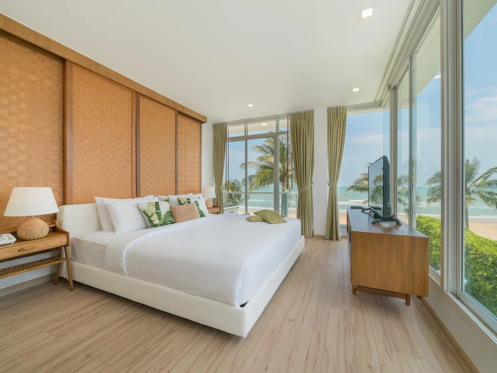 ห้องพักติดทะเลปราณบุรีสไตล์โมเดิร์นพร้อมเตียงขนาดใหญ่ โต๊ะข้างเตียง ทีวีบนขาตั้งไม้ และหน้าต่างบานใหญ่จากพื้นจรดเพดานที่มอบวิวอันงดงามของต้นปาล์มและมหาสมุทร.