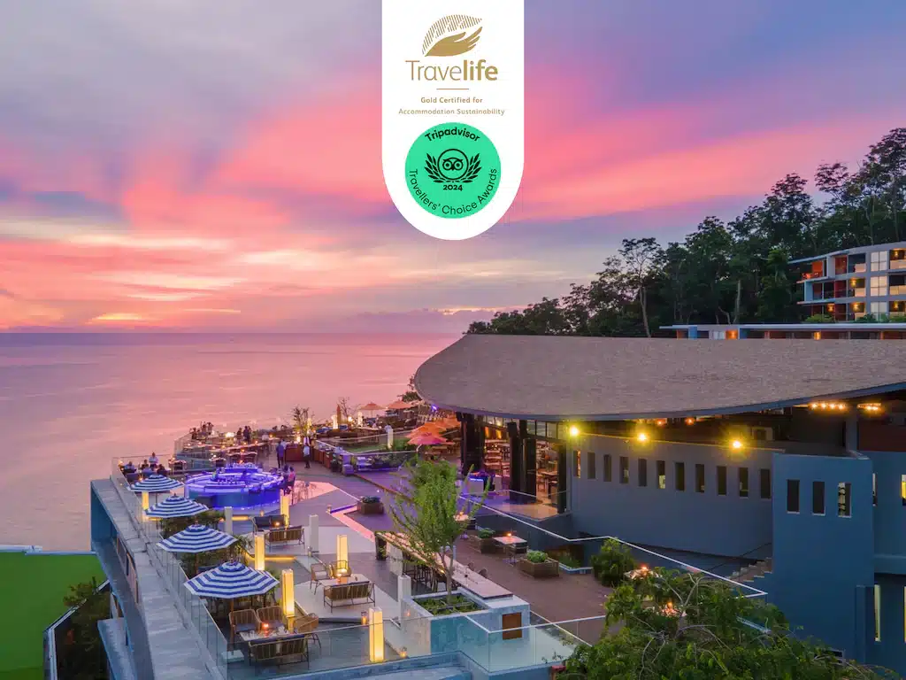รีสอร์ทริมทะเลสมัยใหม่ยามพระอาทิตย์ตกดิน มีที่นั่งกลางแจ้ง พื้นที่สระว่ายน้ำ และวิวทะเลกว้างไกล—สถานที่ท่องเที่ยวป่าตองที่เหมาะอย่างยิ่ง—พร้อมรางวัลด้านความยั่งยืนและรางวัลจาก TripAdvisor ที่แสดงไว้อย่างภาคภูมิใจที่ด้านบนของภาพ.
