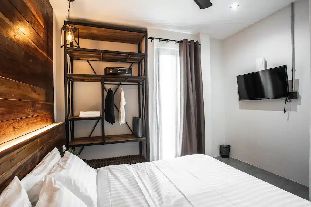 ห้องนอนสไตล์โมเดิร์นพร้อมเตียงคู่ หัวเตียงไม้ ตู้เสื้อผ้าแบบเปิดโล่ง ทีวีติดผนัง และหน้าต่างพร้อมผ้าม่านสีเข้ม ที่พักระนอง เหมาะสำหรับผู้ที่ต้องการที่พักระนองที่มีสไตล์ในระหว่างการเข้าพัก.