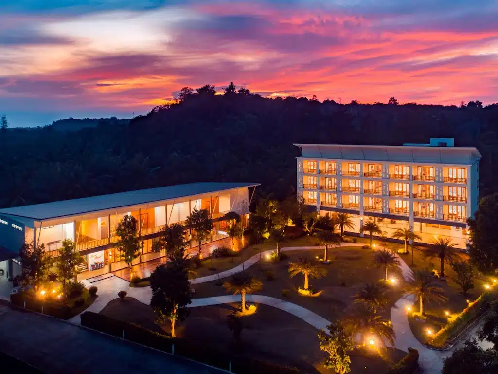 โรงแรมคอมเพล็กซ์สมัยใหม่พร้อมอาคารที่ส่องสว่างและภูมิทัศน์ที่จัดแต่งอย่างสวยงาม ตั้งอยู่ท่ามกลาง ที่เที่ยวชุมพร ต้นไม้และท้องฟ้ายามพระอาทิตย์ตกที่สดใส—สถานที่พักผ่อนที่เชิญชวนใกล้ ชุมพรที่เที่ยว สำหรับนักเดินทางที่มองหาสไตล์และความสะดวกสบาย.