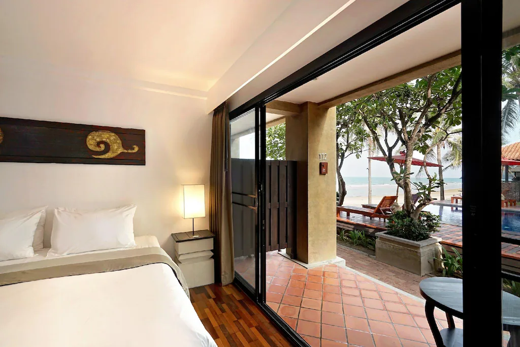 ห้องพักในโรงแรมที่มีเตียงปูผ้าเรียบร้อยและโคมไฟ พร้อมประตูบานเลื่อนกระจกที่เปิดออกสู่ระเบียงซึ่งมีสระว่ายน้ำ ต้นไม้ และวิวทะเล—ที่พักติดทะเลในปราณบุรีที่เหมาะสำหรับการพักผ่อนริมทะเลของคุณ.