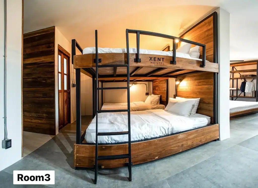 ห้องพักโฮสเทลสมัยใหม่พร้อมเตียงสองชั้นไม้ ผ้าปูที่นอนสีขาว บันไดโลหะ และแสงไฟอบอุ่น ที่พักระนอง เหมาะสำหรับนักเดินทางที่กำลังมองหาที่พักในระนอง พื้นที่นี้ระบุว่าเป็น "ห้อง 3" ที่มุมล่างซ้าย.