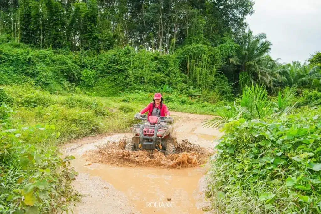 บุคคลที่สวมชุดสีชมพูขี่รถเอทีวีผ่านแอ่งน้ำโคลนบนเส้นทางดินที่ล้อมรอบด้วยพืชพรรณเขียวชอุ่มที่ซีแลนด์แอดเวนเจอร์ พังงา.