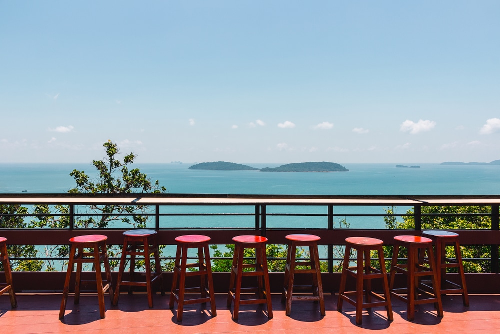 แถวของเก้าอี้บาร์ไม้สีแดงเรียงรายอยู่บนดาดฟ้าที่มองเห็นมหาสมุทรและเกาะไกล ๆ ภายใต้ท้องฟ้าสีฟ้า ชุมพรที่เที่ยว ใส—ภาพที่ชวนให้หลงใหล ณ หนึ่งในสถานที่ท่องเที่ยว ชุมพร ที่ดีที่สุด.