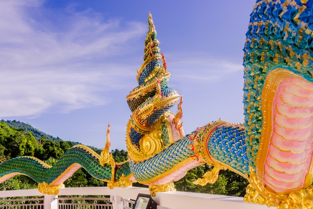 รูปปั้นงูสีสันสดใสและประดับประดาอย่างวิจิตรบรรจง มีเกล็ดที่ซับซ้อนพันรอบราวบันไดของอาคาร ท่ามกลางฉากหลังของต้นไม้และท้องฟ้าสีฟ้า ทำให้เป็นจุดเด่นที่สะดุดตาใน ท่องเที่ยวป่าตอง.