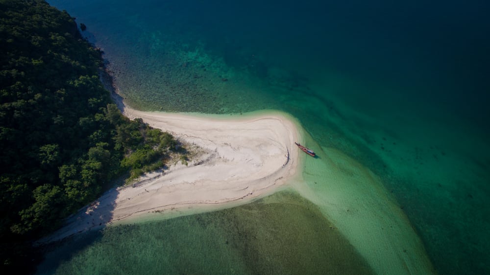 ภาพมุมสูงของชายหาดทรายแคบที่ทอดยาวลงไปในน้ำสีฟ้าเขียวใส ที่เที่ยวชุมพร สะอาด ล้อมรอบด้วยพืชพรรณสีเขียวหนาแน่น มีเรือลำเล็กจอดอยู่ริมฝั่ง—หนึ่งในสถานที่ท่องเที่ยวที่น่าตื่นตาตื่นใจของชุมพร ที่เหมาะสำหรับการหนีไปพักผ่อนอย่างสงบ.